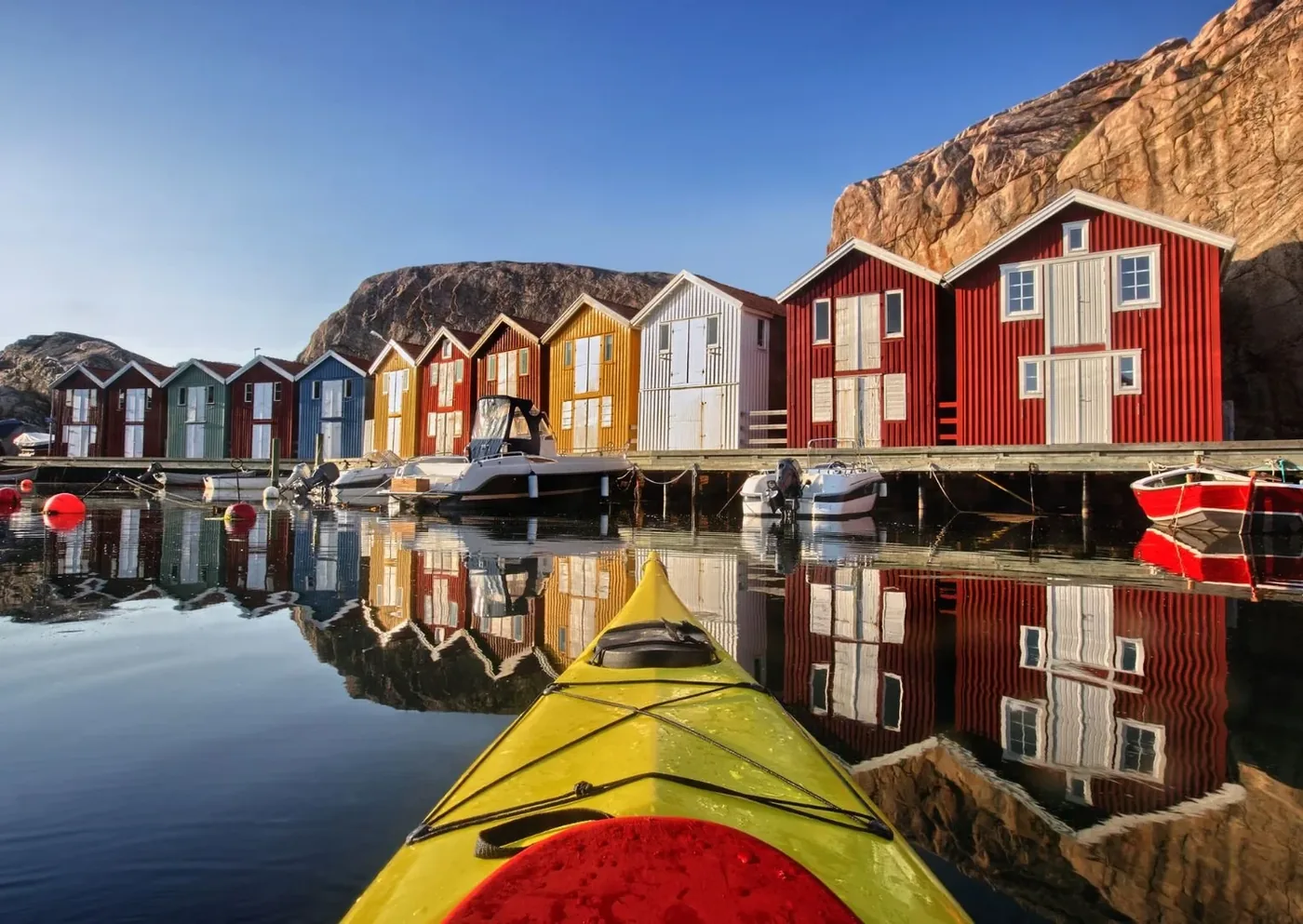 Röda och gula sjöbodar som speglar sig i en svensk hamn, sedda från en kajak